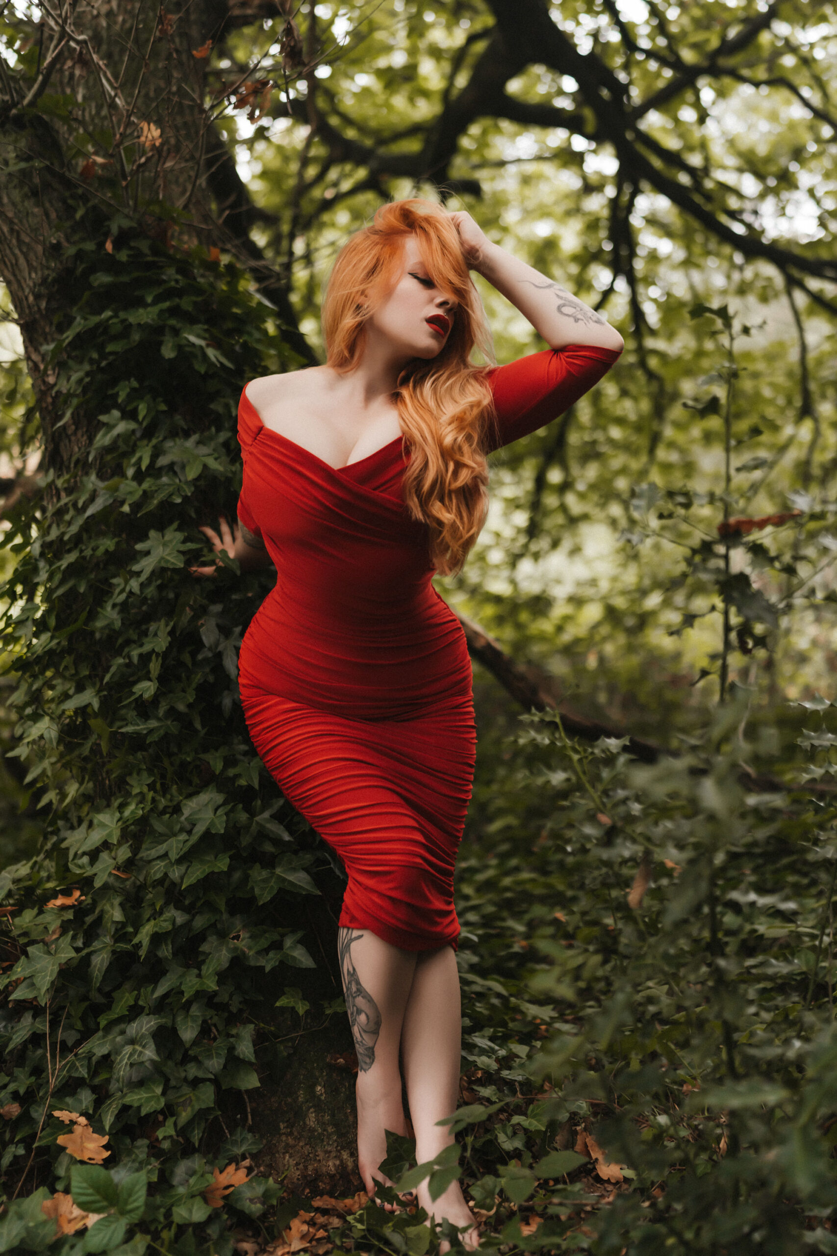 Kinky London escort Amelia Laurent is in a figure-hugging red dress that showcases her hourglass figure. Her red hair cascades down her left shoulder, highlighting her ample cleavage. She is leaning forwards from an ivy-covered tree.